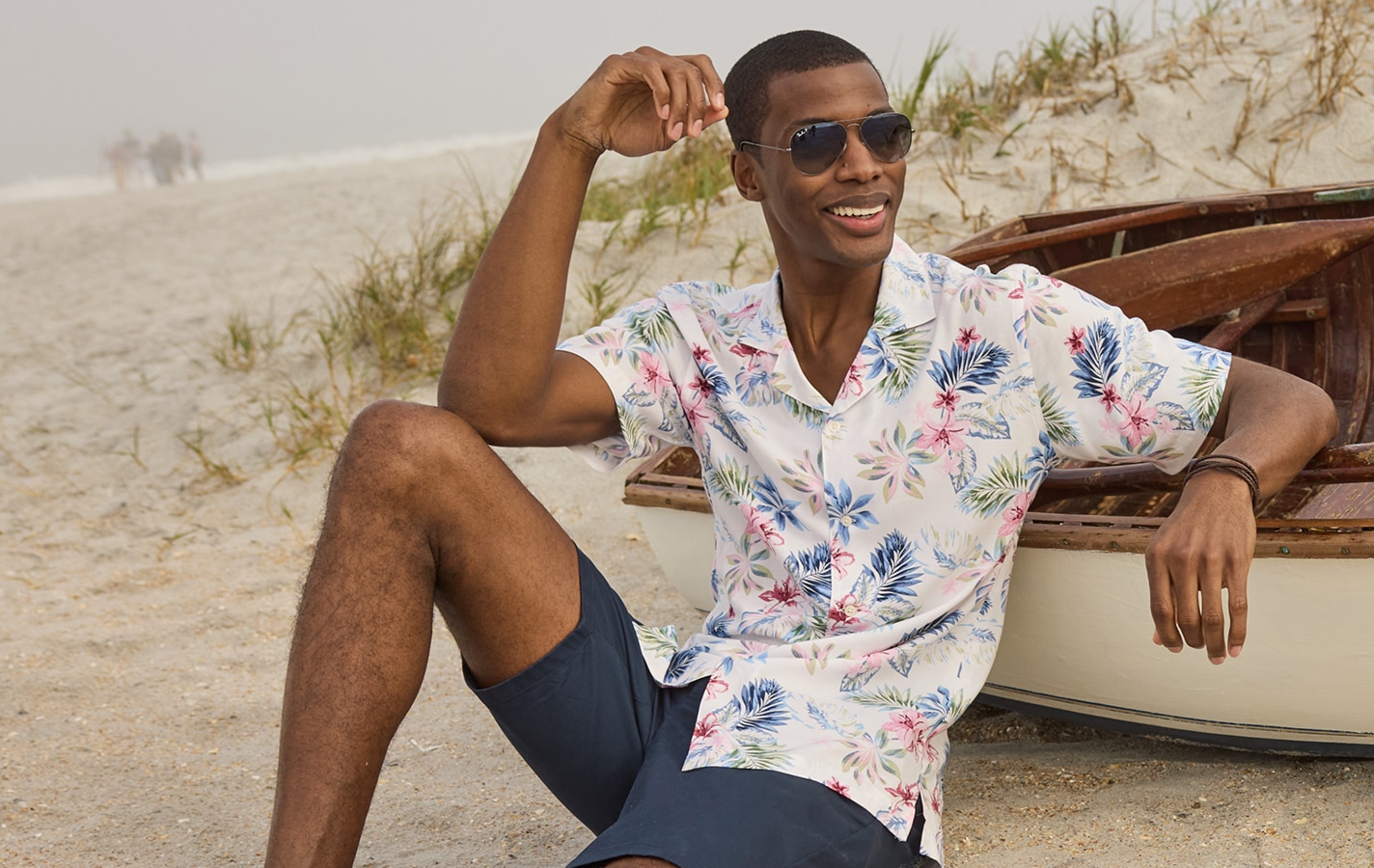 Man in a floral short-sleeve shirt and navy shorts sitting on a beach