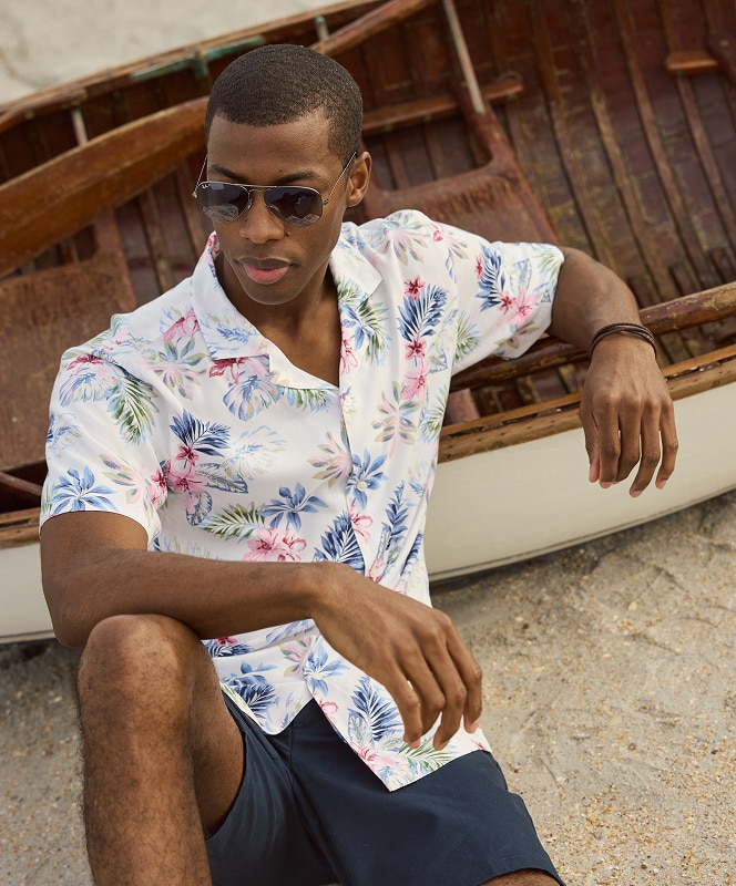 Man in tropical print short-sleeve shirt and navy shorts sitting by a wooden boat on a beach