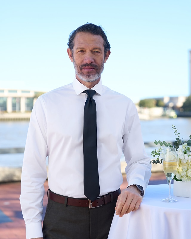 Man in a white dress shirt and black tie at an outdoor waterfront table