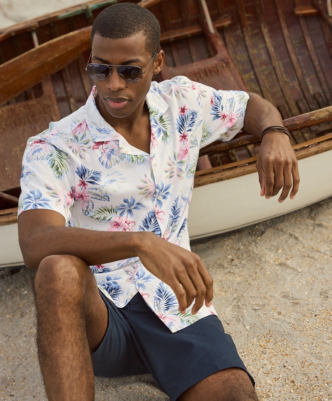 Man wearing a white tropical floral print shirt and navy shorts seated on a beach next to a wooden boat