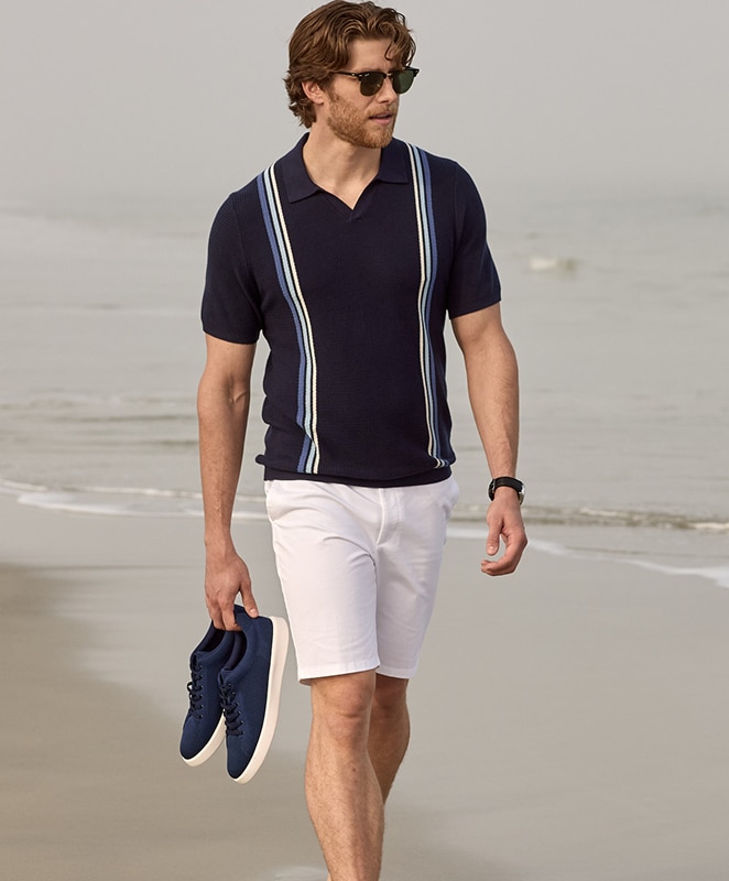 Man in a navy striped polo shirt and white shorts walking on the beach carrying sneakers