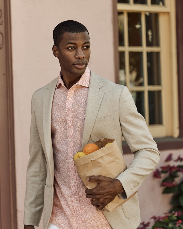 Man in a beige sportcoat and peach patterned dress shirt holding a brown paper bag with fruit