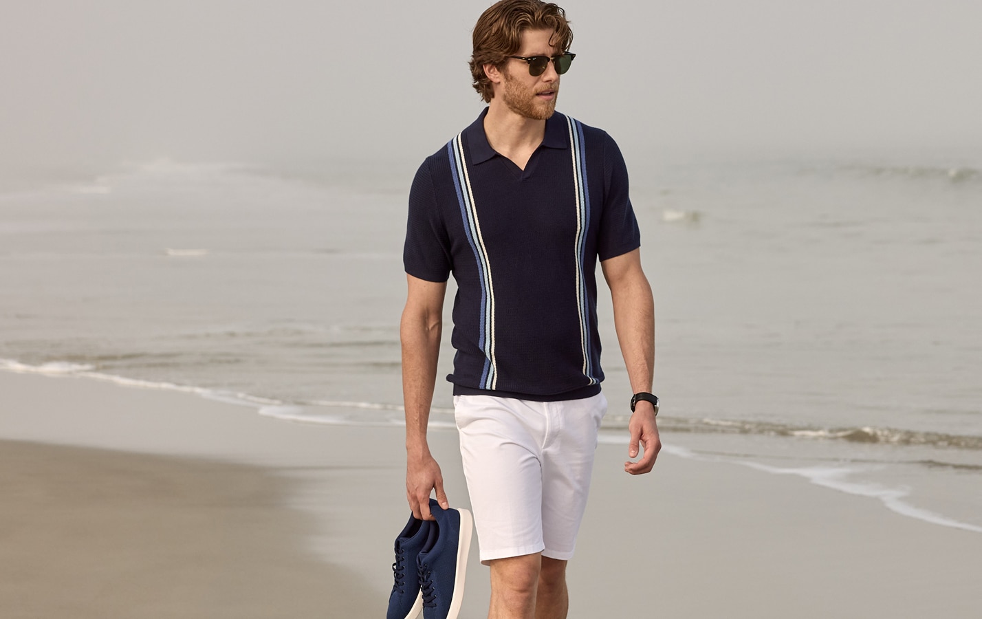 Man in navy striped polo and white shorts walking on the beach