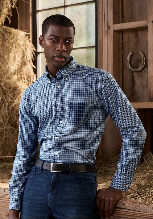 Man wearing a blue and white micro-check casual button-down shirt tucked into dark jeans in a barn setting
