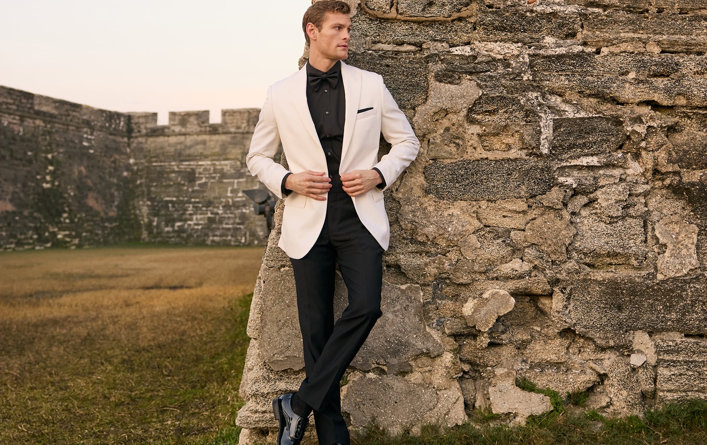 Man in a cream tuxedo jacket, black shirt, and black bow tie leaning against a stone wall