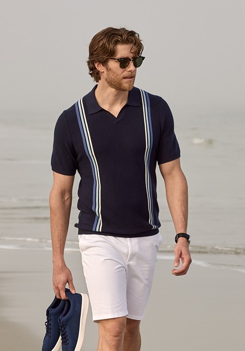 Man in a navy striped polo and white shorts walking on the beach carrying sneakers