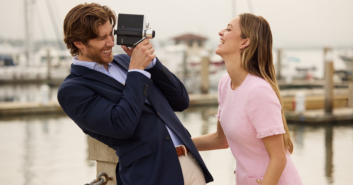 Man in a navy sportcoat filming a woman in a pink dress at a marina