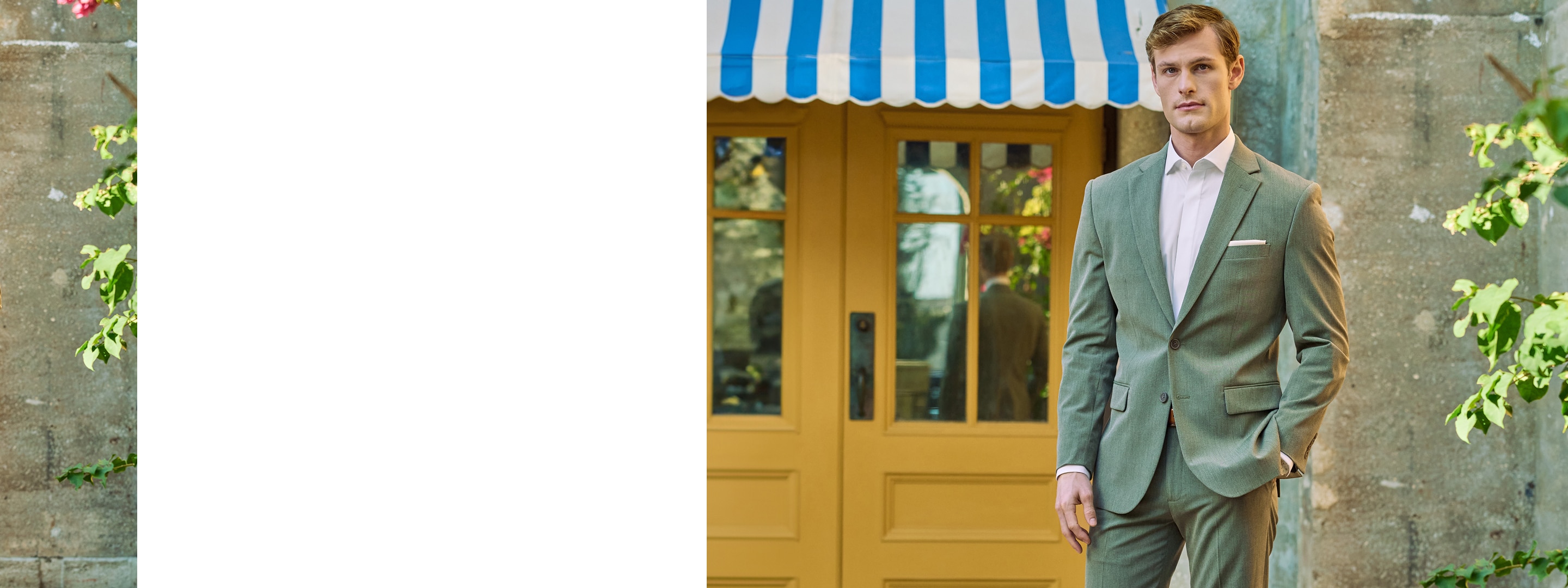 Man in a sage green suit standing in front of yellow doors with a blue and white striped awning