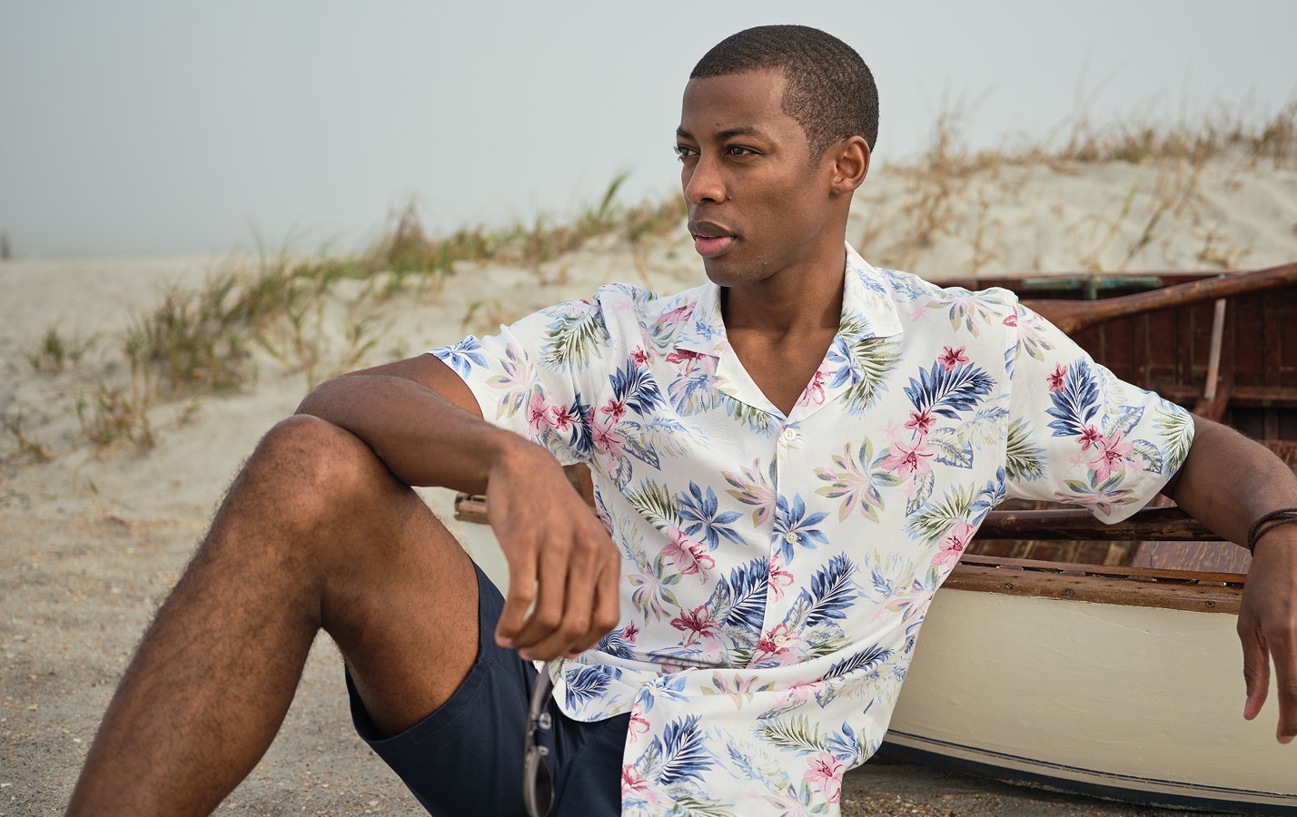 Man in a floral short-sleeve casual shirt sitting on the beach next to a boat