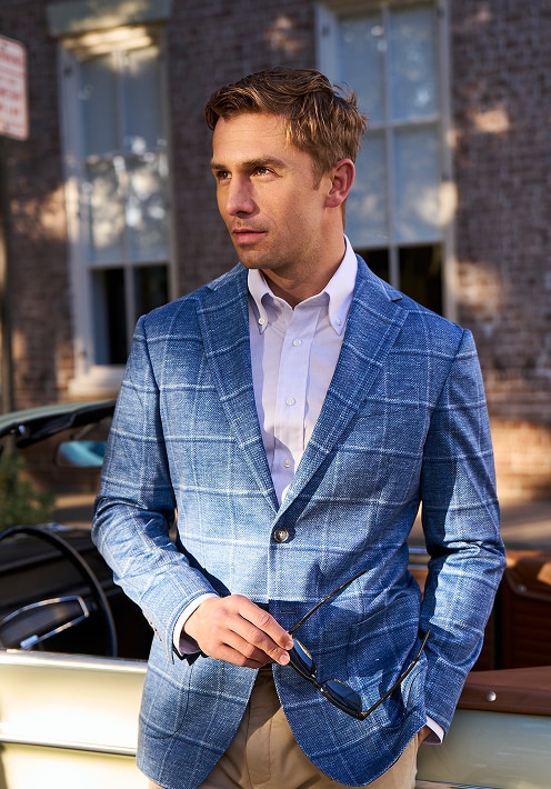 Man in a blue windowpane sportcoat holding sunglasses next to a vintage car