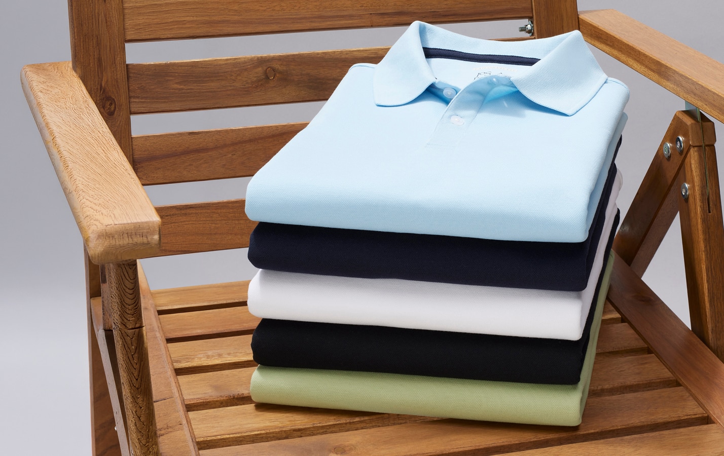 Stack of folded polo shirts in light blue, navy, white, black, and sage on a wooden chair