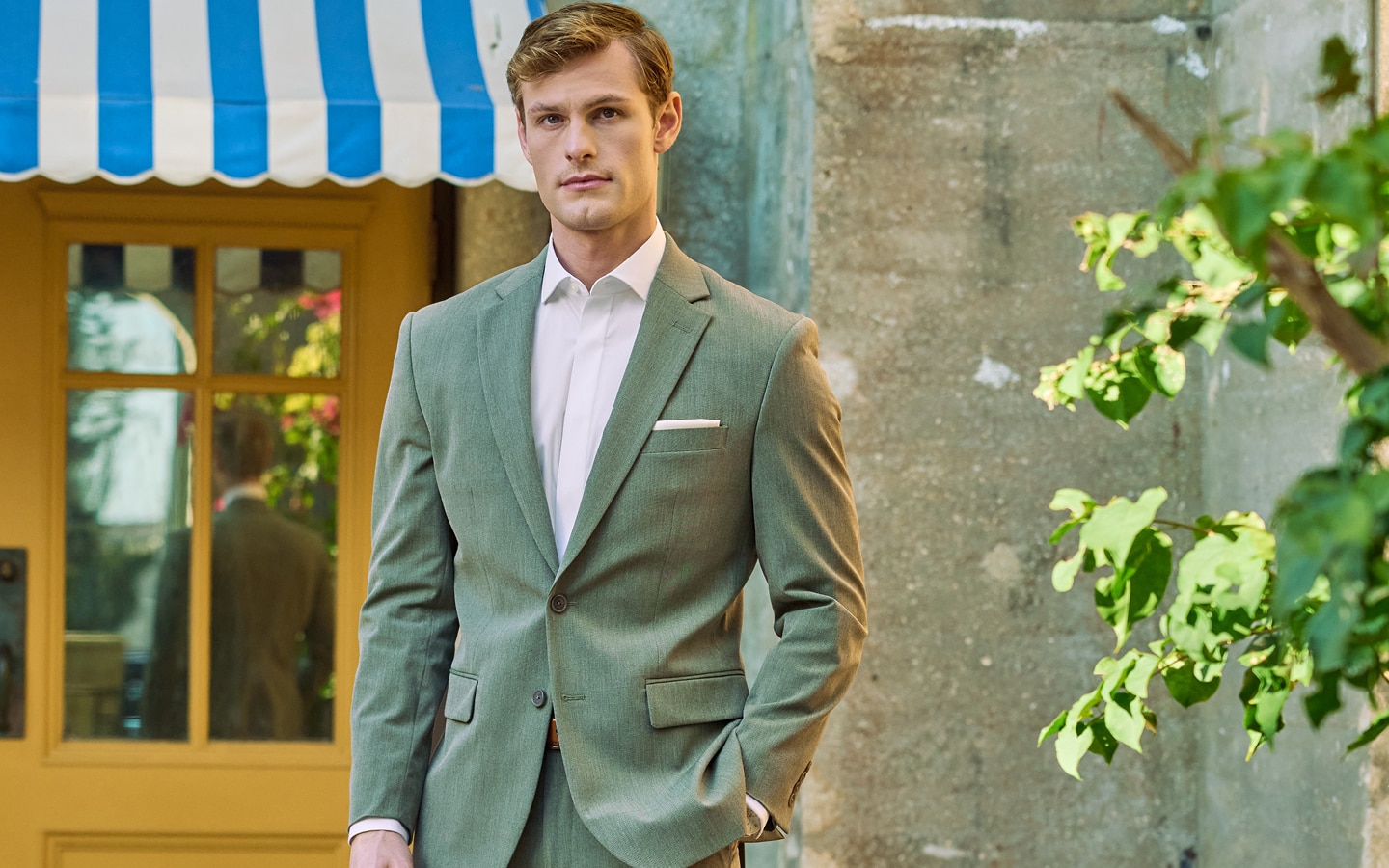 Man in a sage green suit standing in front of yellow doors with a blue and white striped awning