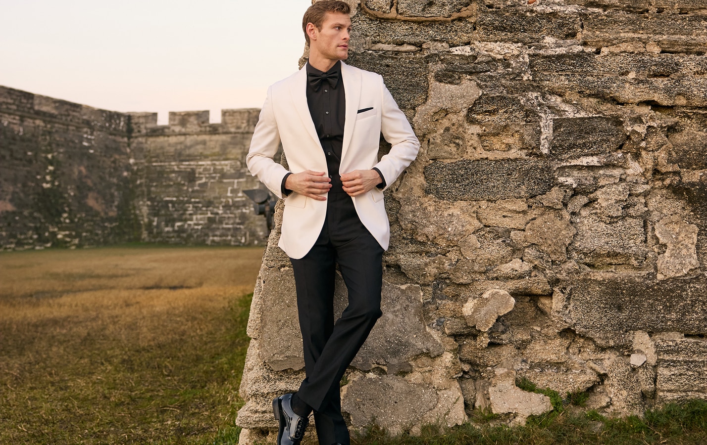 Man in a cream tuxedo jacket, black shirt, and black bow tie leaning against a stone wall
