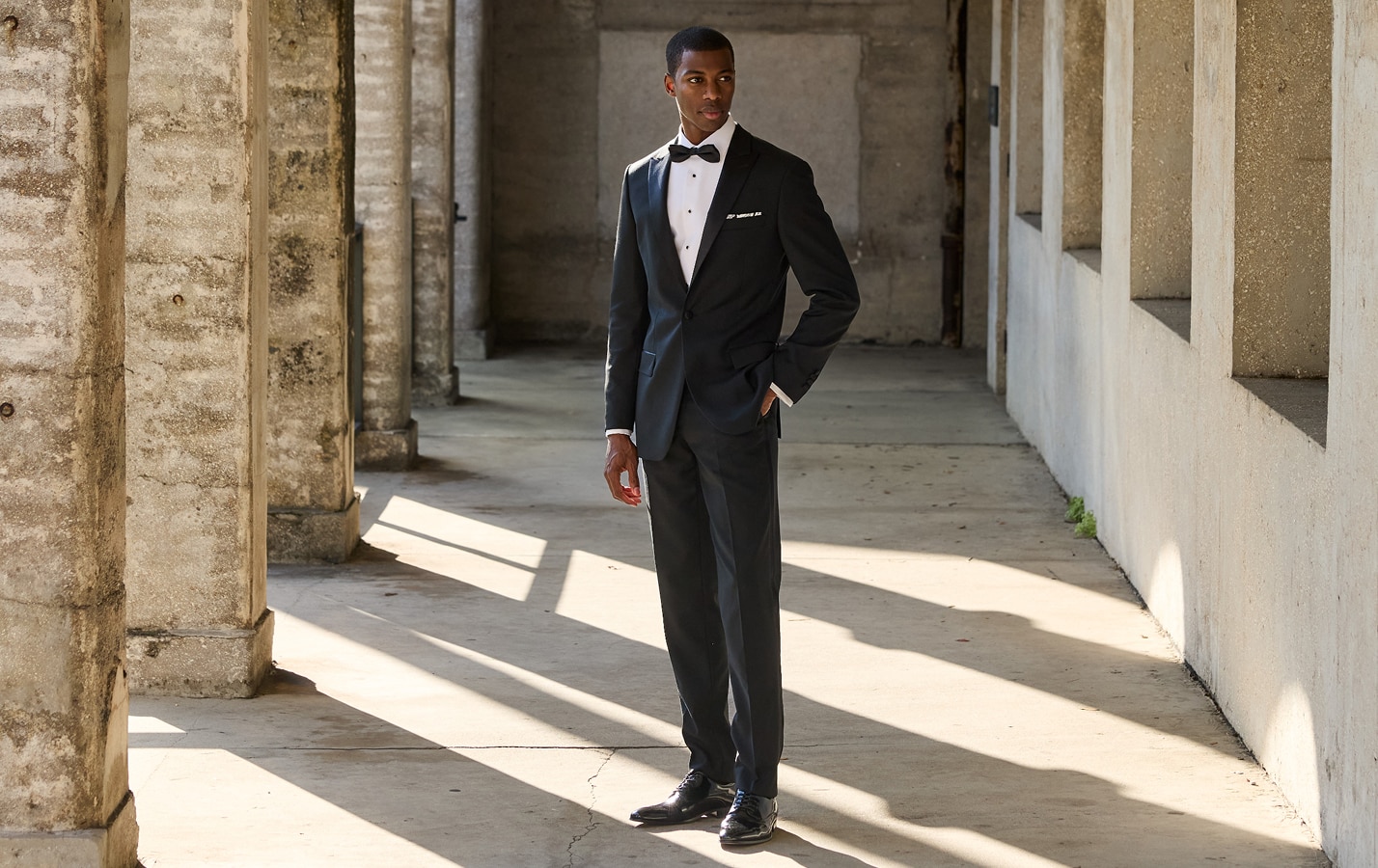 Man in a black tuxedo with bow tie standing in a sunlit stone corridor