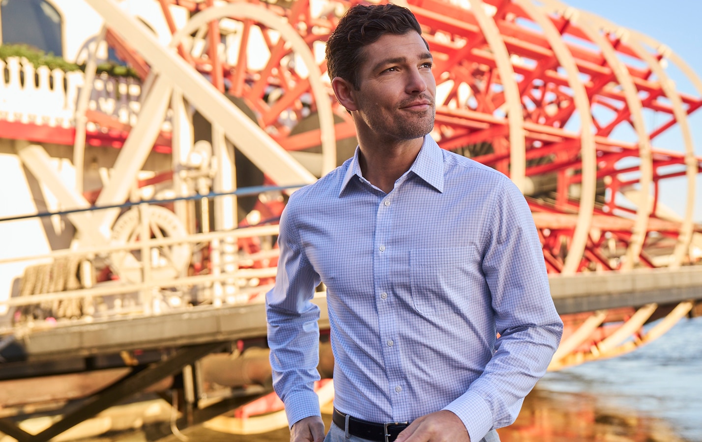 Man wearing a light blue checked dress shirt near a riverboat