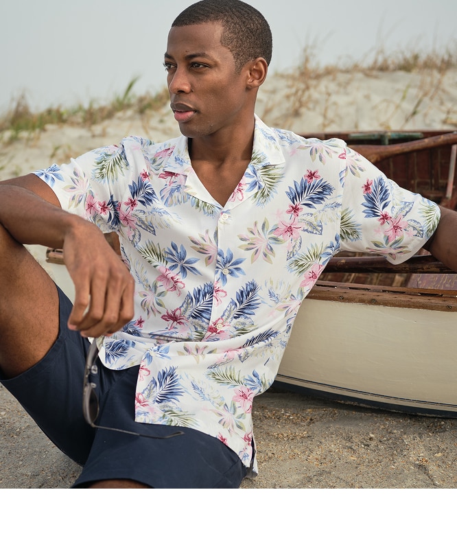 Man wearing a white tropical floral print shirt and navy shorts seated on a beach next to a wooden boat