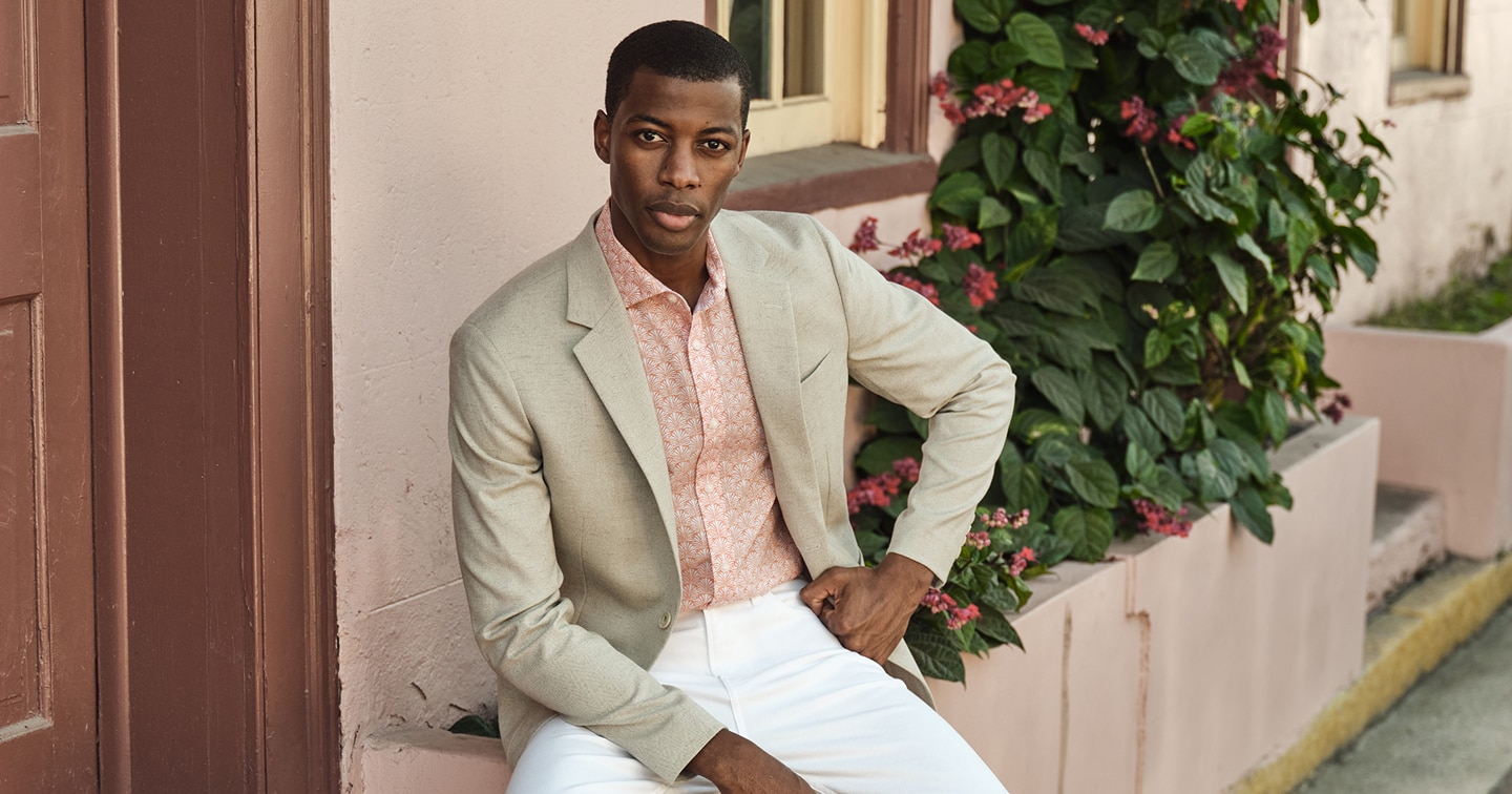 Man in a beige sportcoat and patterned coral shirt sitting outdoors by pink flowers