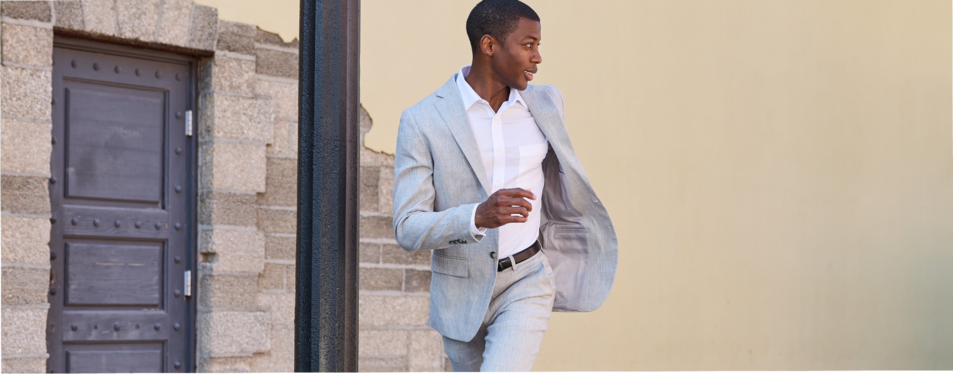 Man in a light grey textured suit walking outdoors past a stone building